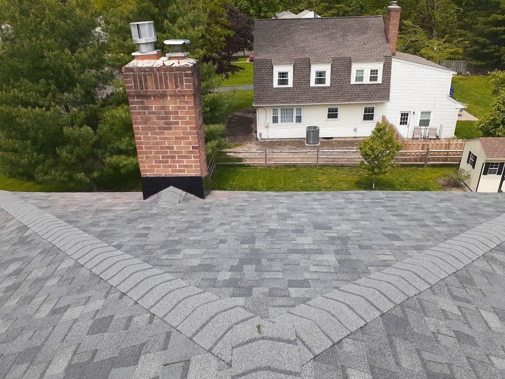 Aerial view of a gray shingle roof with a chimney and a house in the background.